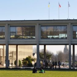 Étudiants profitant du soleil sur l'herbe derrière la galerie vitrée du campus 1 de l'université